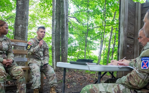 Four U.S. Army soldiers in OCP (Operational Camouflage Pattern) uniforms are engaged in what appears to be a formal evaluation or debriefing session in an outdoor wooded setting. Two soldiers seated on a bench, both wearing '5' numbered patches, face two evaluators seated at a folding table. The soldier on the right side of the table wears a 10th Mountain Division patch, suggesting this may be a training assessment or after-action review.