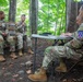 Four U.S. Army soldiers in OCP (Operational Camouflage Pattern) uniforms are engaged in what appears to be a formal evaluation or debriefing session in an outdoor wooded setting. Two soldiers seated on a bench, both wearing '5' numbered patches, face two evaluators seated at a folding table. The soldier on the right side of the table wears a 10th Mountain Division patch, suggesting this may be a training assessment or after-action review.