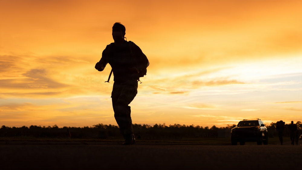A silhouetted figure wearing military-style gear and carrying a large backpack runs across an open field during sunset or sunrise. The dramatic orange and golden sky creates a strong backlit silhouette effect. A vehicle and at least one other person are visible in the background to the right.