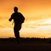 A silhouetted figure wearing military-style gear and carrying a large backpack runs across an open field during sunset or sunrise. The dramatic orange and golden sky creates a strong backlit silhouette effect. A vehicle and at least one other person are visible in the background to the right.