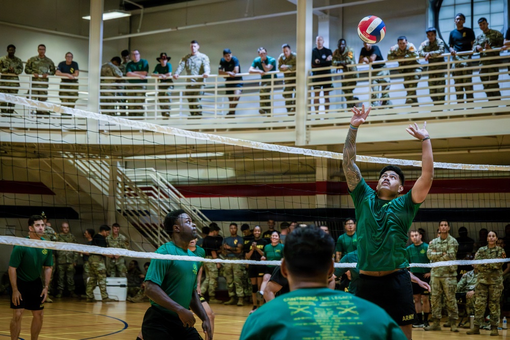 A military volleyball game is taking place inside an indoor gymnasium facility, with soldiers in green team shirts competing against an opposing team. A player in a green shirt is visibly setting or spiking a red, white, and blue volleyball near the net. A large crowd of military personnel in various uniforms watches from both the court level and an upper-level balcony.