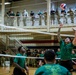A military volleyball game is taking place inside an indoor gymnasium facility, with soldiers in green team shirts competing against an opposing team. A player in a green shirt is visibly setting or spiking a red, white, and blue volleyball near the net. A large crowd of military personnel in various uniforms watches from both the court level and an upper-level balcony.
