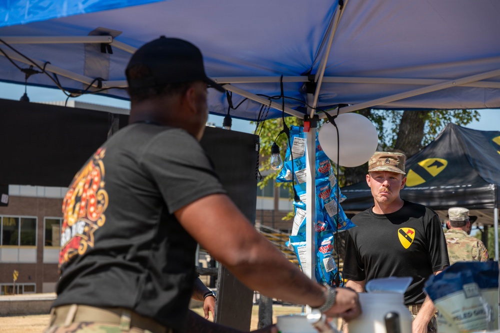 Two individuals wearing black t-shirts with a 1st Cavalry Division patch are working under a blue canopy tent at what appears to be an outdoor military event or cookout. The man in the foreground is out of focus and handling equipment, while the man in the background wearing a camouflage cap looks on attentively. A black tent with the 1st Cavalry Division insignia is visible in the background along with other personnel in military uniforms.