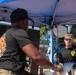 Two individuals wearing black t-shirts with a 1st Cavalry Division patch are working under a blue canopy tent at what appears to be an outdoor military event or cookout. The man in the foreground is out of focus and handling equipment, while the man in the background wearing a camouflage cap looks on attentively. A black tent with the 1st Cavalry Division insignia is visible in the background along with other personnel in military uniforms.