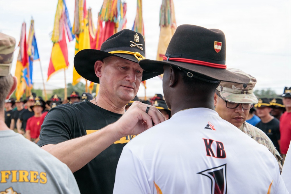 A military ceremony is taking place outdoors, where a senior non-commissioned officer wearing a black cavalry Stetson hat is pinning something onto a soldier wearing a white jersey with 'KB' on the back. Multiple military flags and guidons are visible in the background, along with formations of soldiers in black t-shirts and cavalry hats. The setting appears to be a unit awards or recognition ceremony.