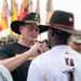 A military ceremony is taking place outdoors, where a senior non-commissioned officer wearing a black cavalry Stetson hat is pinning something onto a soldier wearing a white jersey with 'KB' on the back. Multiple military flags and guidons are visible in the background, along with formations of soldiers in black t-shirts and cavalry hats. The setting appears to be a unit awards or recognition ceremony.