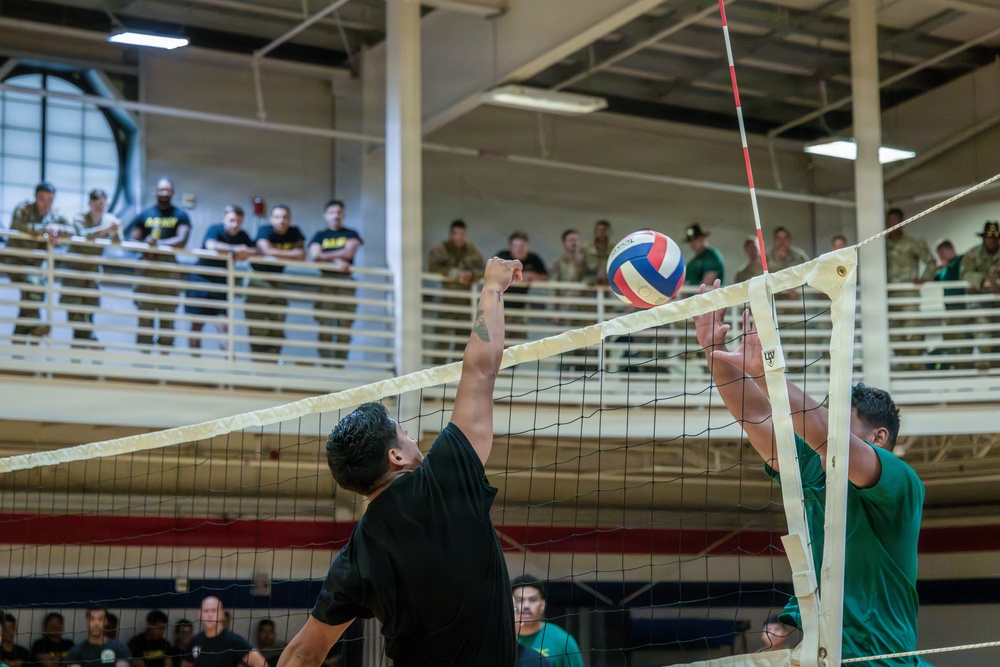 Two players are engaged in a volleyball match at the net inside an indoor gymnasium, with one player in a black shirt attempting to spike or block while the other in a green shirt defends. A large crowd of spectators, many appearing to be in military attire, watches from an upper-level balcony. The setting appears to be a military installation recreational facility.