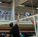 Two players are engaged in a volleyball match at the net inside an indoor gymnasium, with one player in a black shirt attempting to spike or block while the other in a green shirt defends. A large crowd of spectators, many appearing to be in military attire, watches from an upper-level balcony. The setting appears to be a military installation recreational facility.