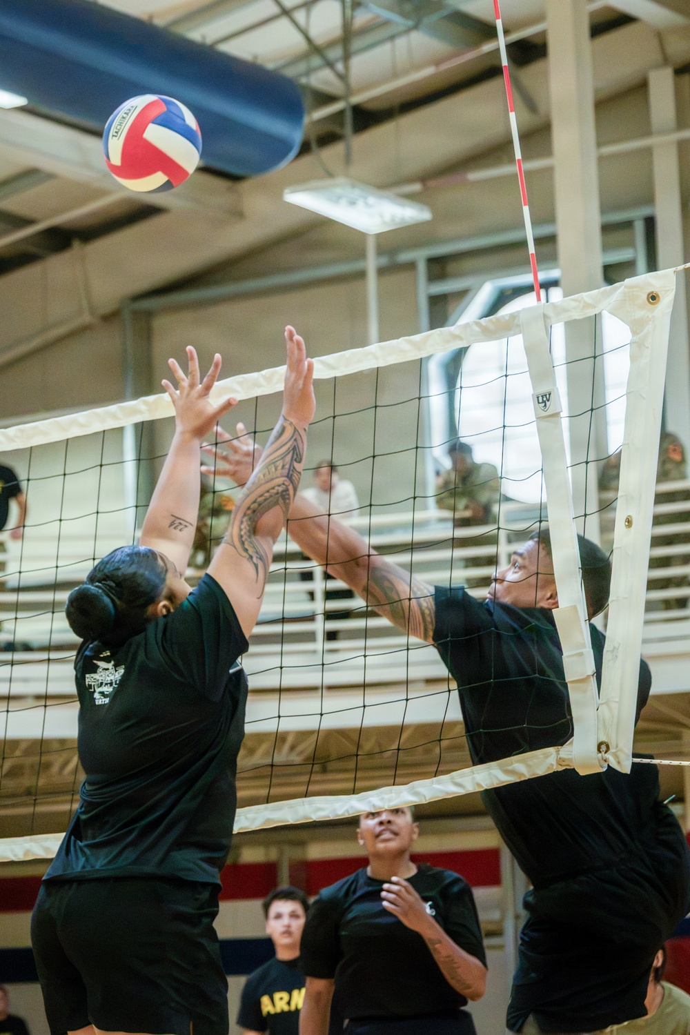 An indoor volleyball match is in progress, with two players in black shirts competing at the net — one attempting a spike while the other reaches up to block. The ball is visible in the upper left, airborne above the net. Spectators and additional players are visible in the background bleachers and court area.