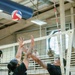 An indoor volleyball match is in progress, with two players in black shirts competing at the net — one attempting a spike while the other reaches up to block. The ball is visible in the upper left, airborne above the net. Spectators and additional players are visible in the background bleachers and court area.