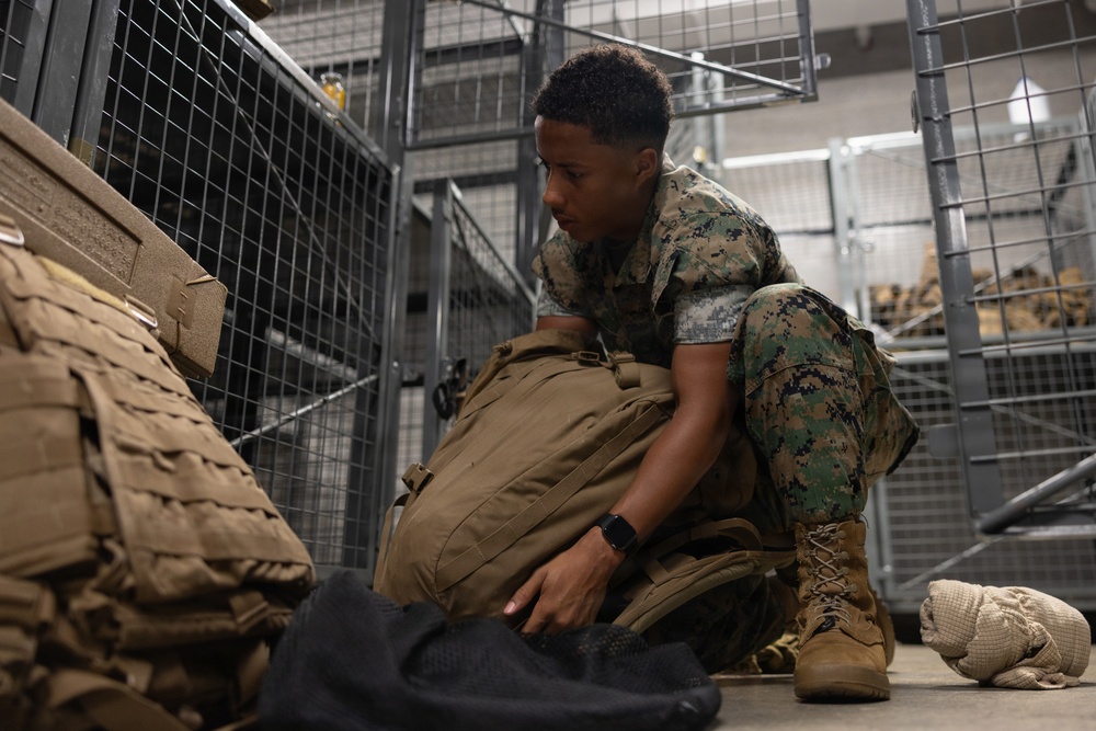 A military service member wearing MARPAT (Marine pattern) digital camouflage uniform is crouched on the floor of what appears to be a military supply or equipment storage facility. The individual is handling a tan/coyote brown tactical backpack among various pieces of military gear. The setting suggests an equipment issue, inventory, or packing operation.
