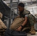 A military service member wearing MARPAT (Marine pattern) digital camouflage uniform is crouched on the floor of what appears to be a military supply or equipment storage facility. The individual is handling a tan/coyote brown tactical backpack among various pieces of military gear. The setting suggests an equipment issue, inventory, or packing operation.
