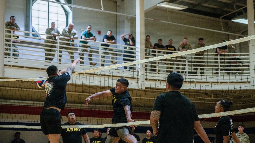 A volleyball game is being played indoors at what appears to be a U.S. Army military facility gymnasium. Players wearing black 'ARMY' t-shirts and shorts compete at the net while a large crowd of spectators, many in military uniforms (ACUs/OCP camouflage), watch from an upper-level balcony. The atmosphere suggests an organized intramural or unit morale event.