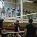 A volleyball game is being played indoors at what appears to be a U.S. Army military facility gymnasium. Players wearing black 'ARMY' t-shirts and shorts compete at the net while a large crowd of spectators, many in military uniforms (ACUs/OCP camouflage), watch from an upper-level balcony. The atmosphere suggests an organized intramural or unit morale event.