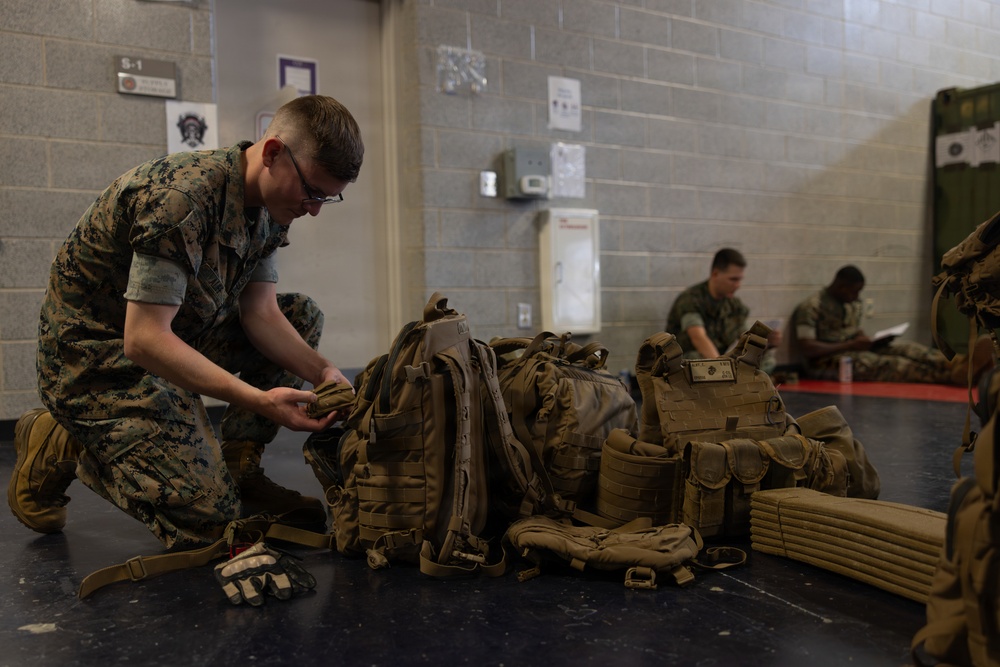 A U.S. Marine in MARPAT digital camouflage uniform is kneeling on a dark floor, inspecting or organizing a pile of tactical gear including backpacks and plate carriers. The setting appears to be an indoor military facility or gymnasium, with cinder block walls and posted notices visible. Additional personnel in the background are seated and appear to be reviewing documents.