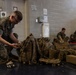 A U.S. Marine in MARPAT digital camouflage uniform is kneeling on a dark floor, inspecting or organizing a pile of tactical gear including backpacks and plate carriers. The setting appears to be an indoor military facility or gymnasium, with cinder block walls and posted notices visible. Additional personnel in the background are seated and appear to be reviewing documents.