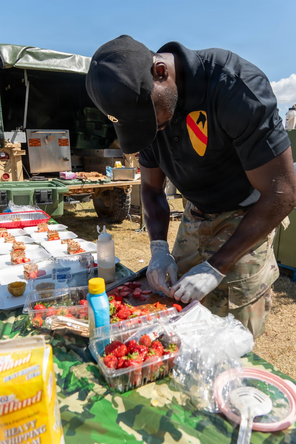 A soldier wearing a 1st Cavalry Division patch on a black polo shirt prepares food at an outdoor military field kitchen setup. He is wearing disposable gloves and arranging strawberries on a tray, with various dessert items and ingredients spread across a camouflage-covered table. The setting appears to be a military culinary competition or field feeding event.
