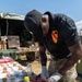A soldier wearing a 1st Cavalry Division patch on a black polo shirt prepares food at an outdoor military field kitchen setup. He is wearing disposable gloves and arranging strawberries on a tray, with various dessert items and ingredients spread across a camouflage-covered table. The setting appears to be a military culinary competition or field feeding event.