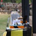 A military ceremony featuring a row of decorated sheet cakes displayed on tables covered with black tablecloths. The cakes are decorated in black, white, and gold/orange colors, consistent with U.S. Army unit colors, and military sabers are placed alongside them. A large crowd of uniformed personnel is visible in the blurred background, suggesting a formal unit or Army birthday celebration.