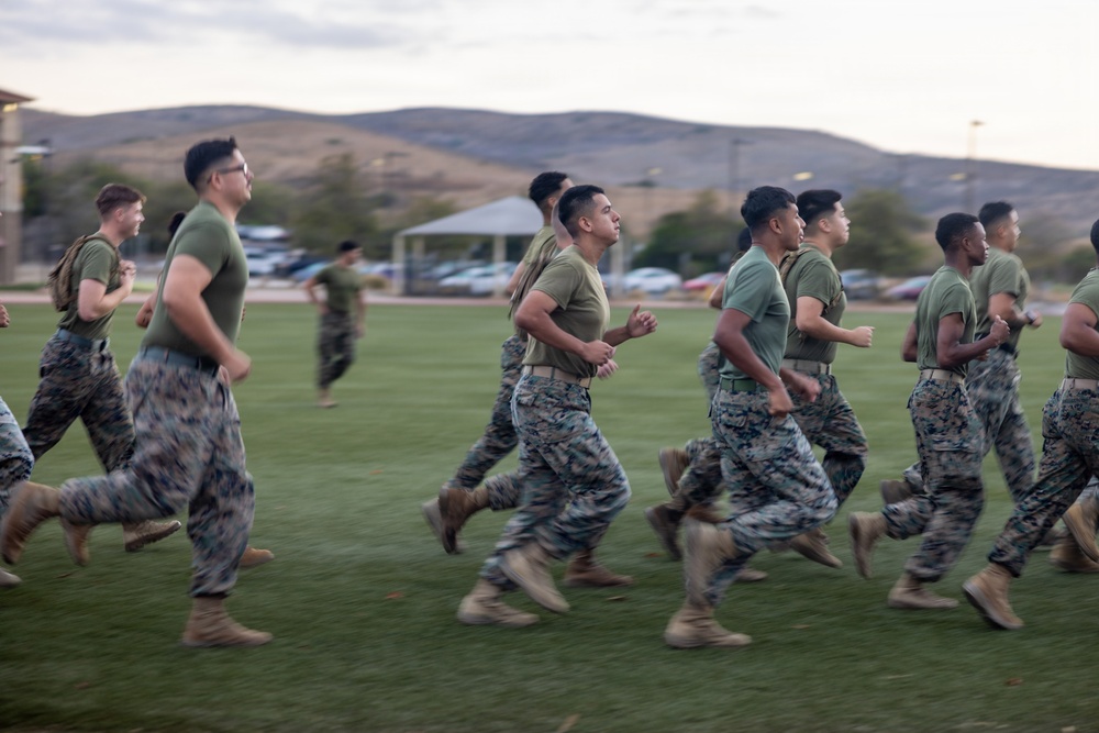 A group of military personnel in uniform are conducting a formation run across a grass field, likely during physical training (PT). They are wearing matching olive green t-shirts and Marine Corps MARPAT camouflage trousers with tan combat boots. The setting appears to be a military installation with a parking lot, pavilion structure, and rolling hills visible in the background.