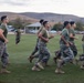 A group of military personnel in uniform are conducting a formation run across a grass field, likely during physical training (PT). They are wearing matching olive green t-shirts and Marine Corps MARPAT camouflage trousers with tan combat boots. The setting appears to be a military installation with a parking lot, pavilion structure, and rolling hills visible in the background.