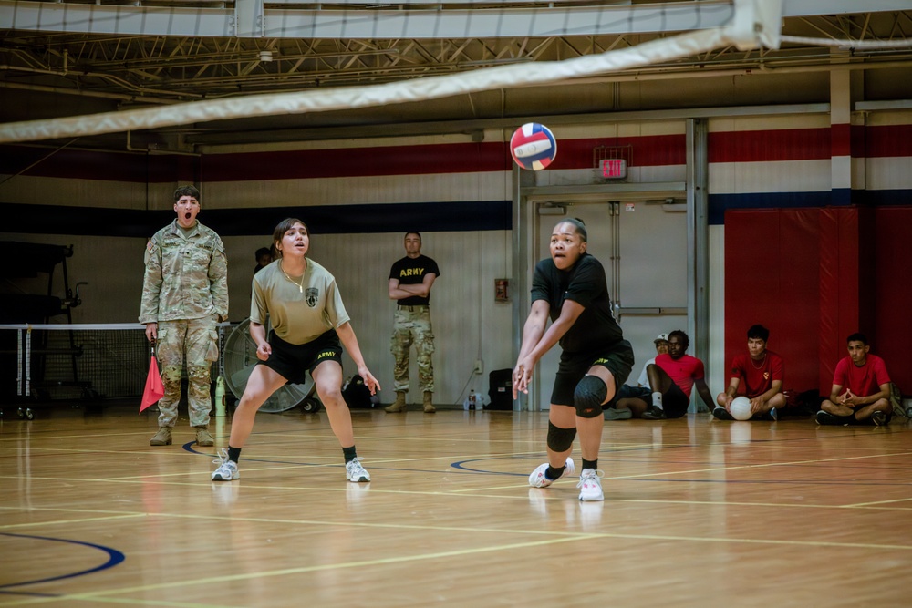 A volleyball game is being played indoors in a military gymnasium, with two female soldiers actively engaged on the court. One player in black Army PT gear is executing a forearm pass (bump) while her teammate in olive Army PT gear stands ready nearby. Spectators in red shirts sit along the wall, and a soldier in full camouflage uniform observes from the sideline.