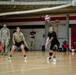 A volleyball game is being played indoors in a military gymnasium, with two female soldiers actively engaged on the court. One player in black Army PT gear is executing a forearm pass (bump) while her teammate in olive Army PT gear stands ready nearby. Spectators in red shirts sit along the wall, and a soldier in full camouflage uniform observes from the sideline.