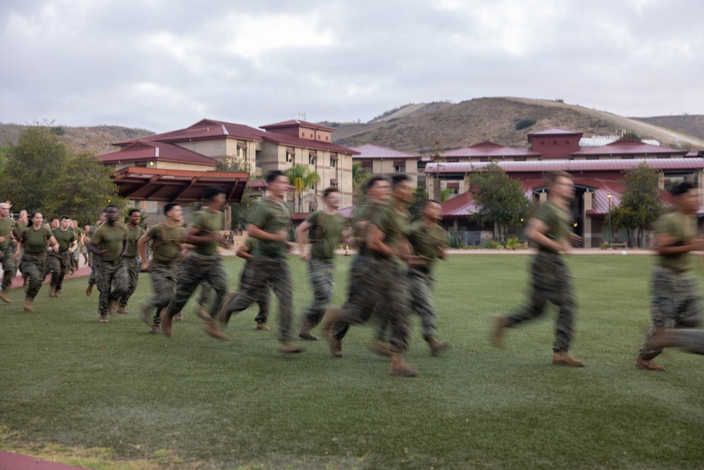 A large group of military personnel in olive green t-shirts and camouflage trousers are running across a grass field at what appears to be a military installation. The motion blur in the image conveys the speed and energy of the formation run. The background features multi-story buildings with red roofs, palm trees, and rolling hills under an overcast sky.