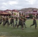 A large group of military personnel in olive green t-shirts and camouflage trousers are running across a grass field at what appears to be a military installation. The motion blur in the image conveys the speed and energy of the formation run. The background features multi-story buildings with red roofs, palm trees, and rolling hills under an overcast sky.