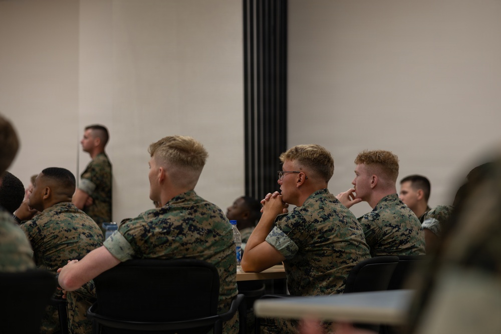 A group of military personnel in camouflage uniforms are seated at desks in what appears to be a classroom or briefing room setting. The individuals are attentively focused toward the front of the room, suggesting a lecture, briefing, or training session is in progress. One service member is visible standing near the back wall.