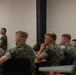 A group of military personnel in camouflage uniforms are seated at desks in what appears to be a classroom or briefing room setting. The individuals are attentively focused toward the front of the room, suggesting a lecture, briefing, or training session is in progress. One service member is visible standing near the back wall.