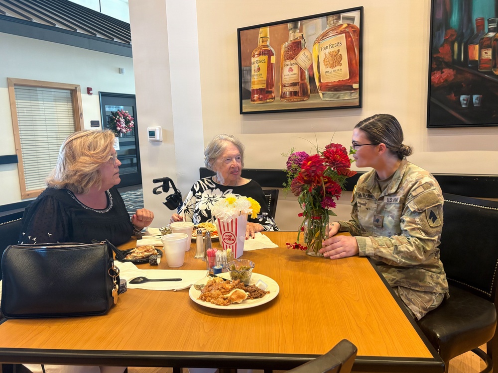 Three women are seated at a restaurant table engaged in conversation. One woman is wearing a U.S. Army camouflage uniform with a name tape reading 'McKENNEY', while the other two are in civilian clothing. The table has food, flowers, a popcorn container, and condiments on it, with Four Roses bourbon artwork visible on the wall behind them.