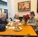 Three women are seated at a restaurant table engaged in conversation. One woman is wearing a U.S. Army camouflage uniform with a name tape reading 'McKENNEY', while the other two are in civilian clothing. The table has food, flowers, a popcorn container, and condiments on it, with Four Roses bourbon artwork visible on the wall behind them.