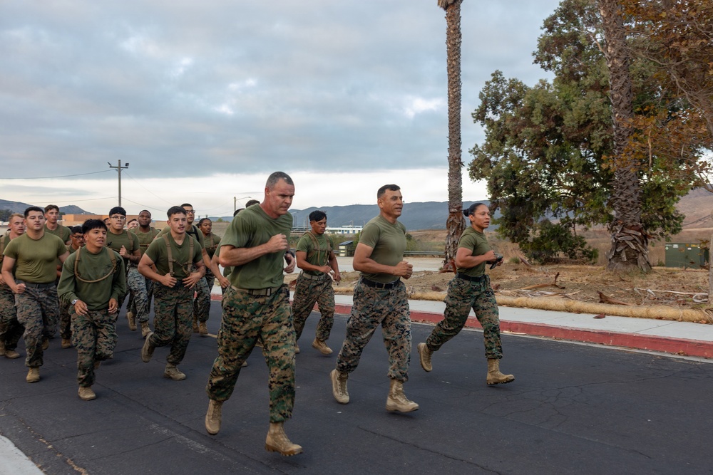 A large group of military personnel in U.S. Marine Corps uniforms are conducting a formation run on a paved road at what appears to be a military installation. The group is wearing matching olive green t-shirts with Marine Corps camouflage trousers and tan combat boots. The setting appears to be a base in a semi-arid region, likely in California based on the terrain and vegetation.