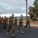 A large group of military personnel in U.S. Marine Corps uniforms are conducting a formation run on a paved road at what appears to be a military installation. The group is wearing matching olive green t-shirts with Marine Corps camouflage trousers and tan combat boots. The setting appears to be a base in a semi-arid region, likely in California based on the terrain and vegetation.