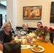 Three women are seated together at a dining table, smiling for the photo. The group includes an older woman in a floral top, a middle-aged woman in black, and a younger woman in U.S. Army military uniform. They appear to be sharing a meal in what looks like a restaurant or dining facility decorated with bourbon-themed artwork.