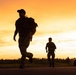 A dramatic silhouette photograph taken at sunset or sunrise showing military or tactical personnel running along a road. The warm orange and yellow sky creates a strong backlit effect, rendering the figures as dark silhouettes. At least three individuals and one vehicle are visible in the scene.