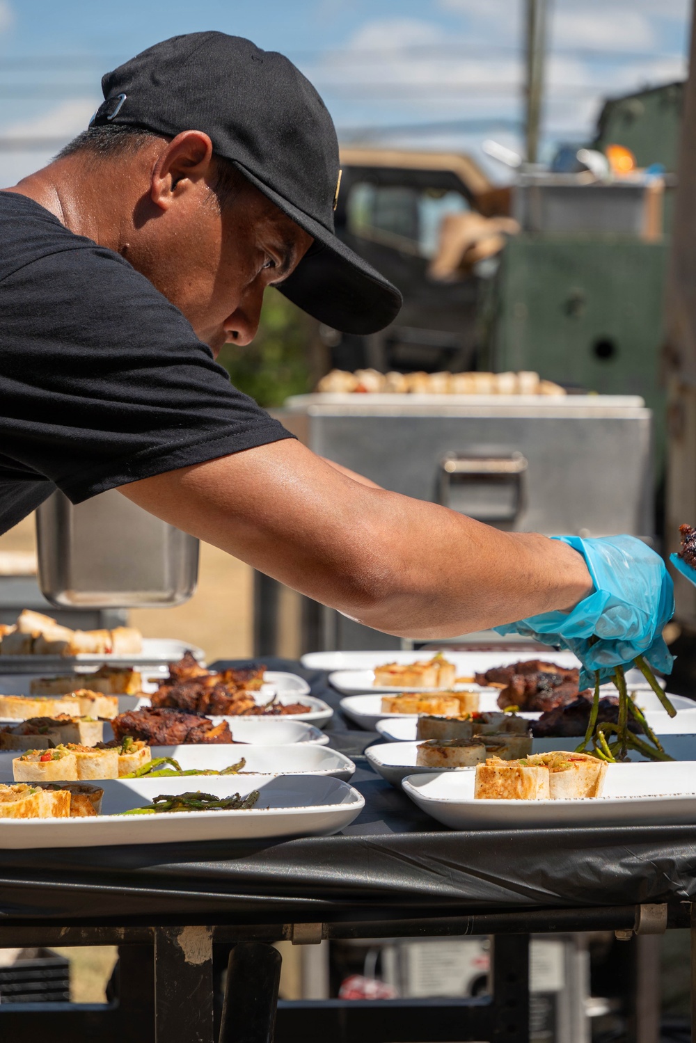 A food service worker wearing a black cap, black t-shirt, and blue latex gloves is carefully plating multiple dishes on white rectangular plates arranged on a black-covered outdoor table. The scene appears to be an outdoor catering or food competition event, with numerous identical plates being prepared simultaneously. In the background, metal food warmers/containers and what appears to be a military-style vehicle are visible.
