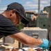 A food service worker wearing a black cap, black t-shirt, and blue latex gloves is carefully plating multiple dishes on white rectangular plates arranged on a black-covered outdoor table. The scene appears to be an outdoor catering or food competition event, with numerous identical plates being prepared simultaneously. In the background, metal food warmers/containers and what appears to be a military-style vehicle are visible.