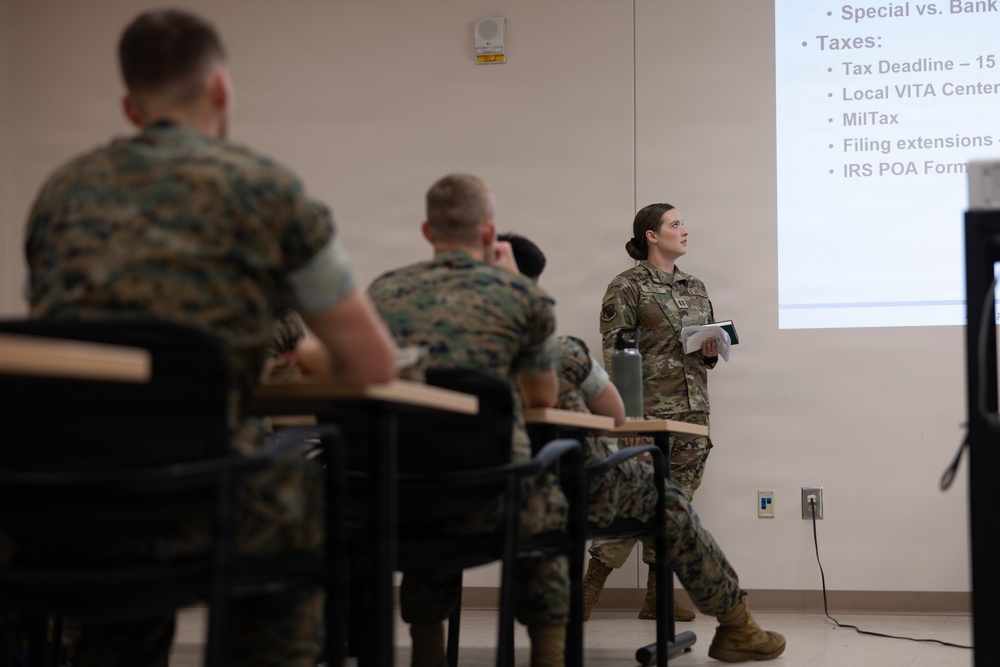 A military briefing or training session is taking place in a classroom setting. A female service member in Army Combat Uniform (ACU/OCP pattern) is presenting a financial/tax briefing to an audience of Marines in MARPAT camouflage uniforms. The projected presentation covers tax-related topics including VITA centers, MilTax, filing extensions, and IRS POA forms.