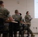 A military briefing or training session is taking place in a classroom setting. A female service member in Army Combat Uniform (ACU/OCP pattern) is presenting a financial/tax briefing to an audience of Marines in MARPAT camouflage uniforms. The projected presentation covers tax-related topics including VITA centers, MilTax, filing extensions, and IRS POA forms.