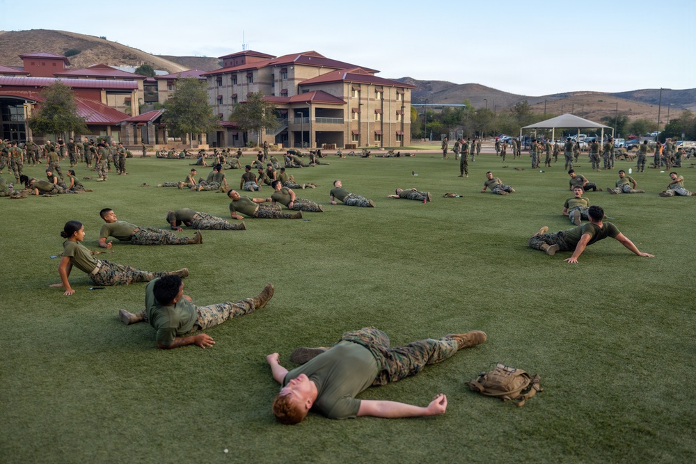A large group of military personnel in camouflage uniforms and olive green t-shirts are conducting physical training on a grass field. They appear to be performing stretching or cool-down exercises, with many lying prone or in cobra/back-extension positions. The setting appears to be a military installation with multi-story barracks buildings and rolling hills visible in the background.