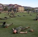 A large group of military personnel in camouflage uniforms and olive green t-shirts are conducting physical training on a grass field. They appear to be performing stretching or cool-down exercises, with many lying prone or in cobra/back-extension positions. The setting appears to be a military installation with multi-story barracks buildings and rolling hills visible in the background.