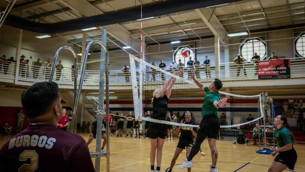 A military volleyball tournament is taking place inside a gymnasium, with players in Army PT uniforms and civilian athletic wear competing across multiple courts. Spectators in military uniforms observe from an upper-level balcony and ground level. A banner reading 'OMNI - A Helping Hand for Military Life' is visible in the background.