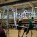 A military volleyball tournament is taking place inside a gymnasium, with players in Army PT uniforms and civilian athletic wear competing across multiple courts. Spectators in military uniforms observe from an upper-level balcony and ground level. A banner reading 'OMNI - A Helping Hand for Military Life' is visible in the background.