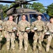 Seven U.S. Army soldiers in OCP (Operational Camouflage Pattern) uniforms pose for a group photo in front of a UH-60 Black Hawk helicopter on a grassy field. Each soldier is holding a numbered military helmet, suggesting participation in a training exercise or airborne/air assault operation. The setting appears to be a military training area with trees visible in the background.