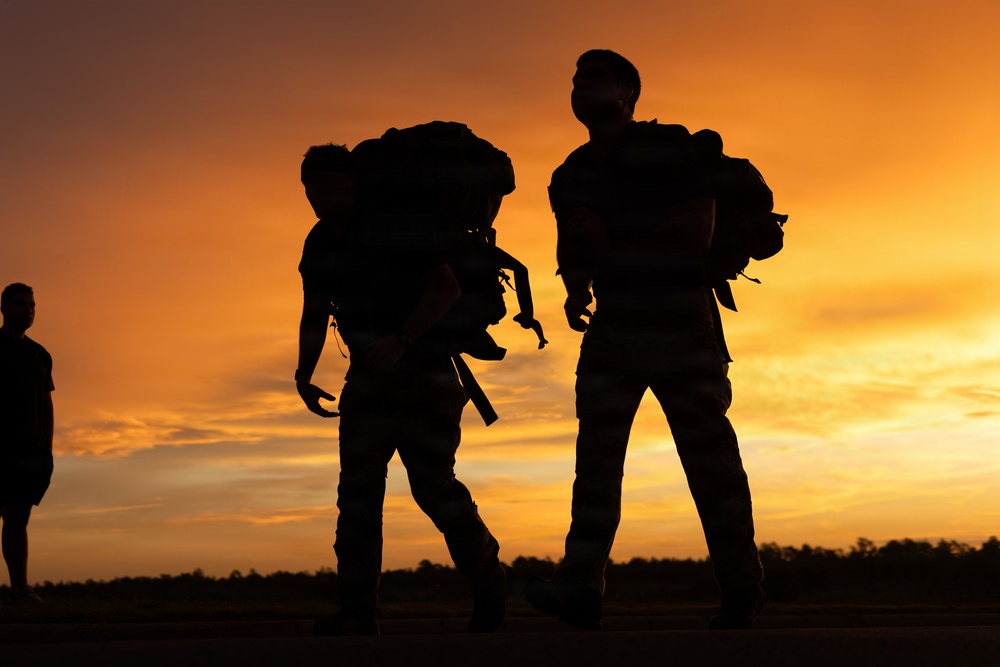 A dramatic silhouette photograph taken at sunset showing multiple individuals carrying large backpacks on what appears to be a flat open surface, likely a tarmac or field. The warm orange and amber tones of the sky create a striking backlit effect, rendering the subjects as dark silhouettes. The large packs and overall posture of the figures suggest military, tactical, or heavy outdoor expedition activity.