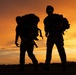 A dramatic silhouette photograph taken at sunset showing multiple individuals carrying large backpacks on what appears to be a flat open surface, likely a tarmac or field. The warm orange and amber tones of the sky create a striking backlit effect, rendering the subjects as dark silhouettes. The large packs and overall posture of the figures suggest military, tactical, or heavy outdoor expedition activity.