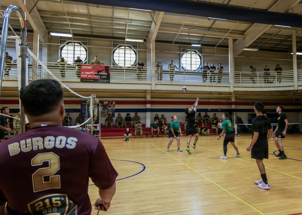 A military volleyball tournament is taking place inside a gymnasium, with players in mixed uniforms competing on a hardwood court. Spectators in military uniforms observe from both ground-level bleachers and an upper-level mezzanine balcony. A banner for 'Omni - A Helping Hand for Military Life' is visible on the upper level railing.