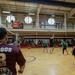 A military volleyball tournament is taking place inside a gymnasium, with players in mixed uniforms competing on a hardwood court. Spectators in military uniforms observe from both ground-level bleachers and an upper-level mezzanine balcony. A banner for 'Omni - A Helping Hand for Military Life' is visible on the upper level railing.